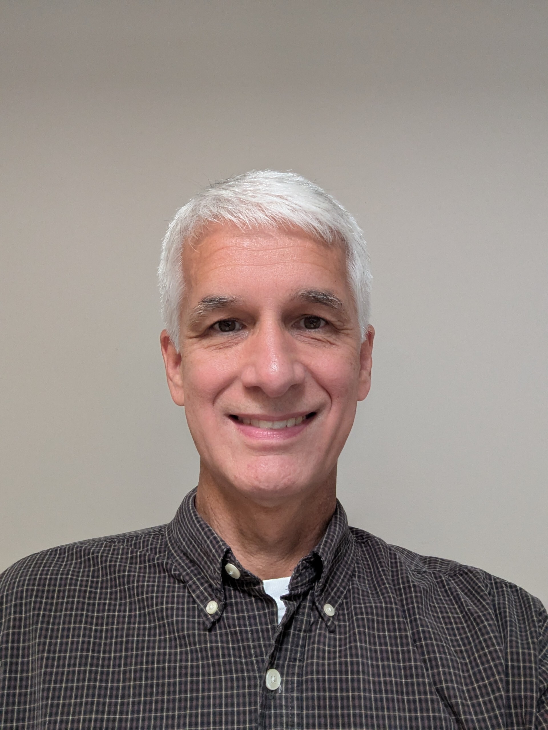 Smiling person with short white hair wearing a dark plaid button-down shirt, neutral gray background