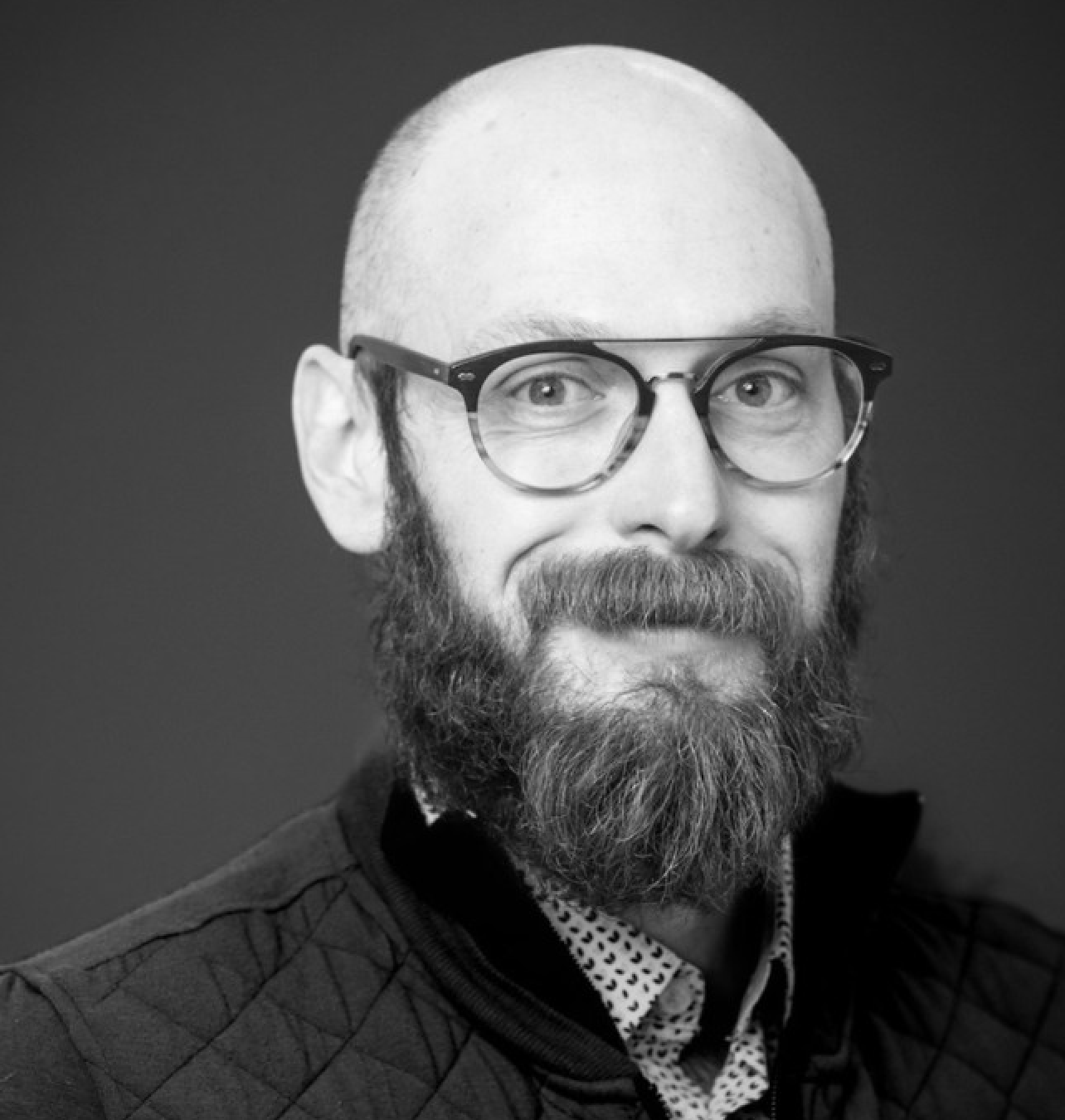 Black-and-white portrait of a person with a shaved head, beard, and round-frame glasses