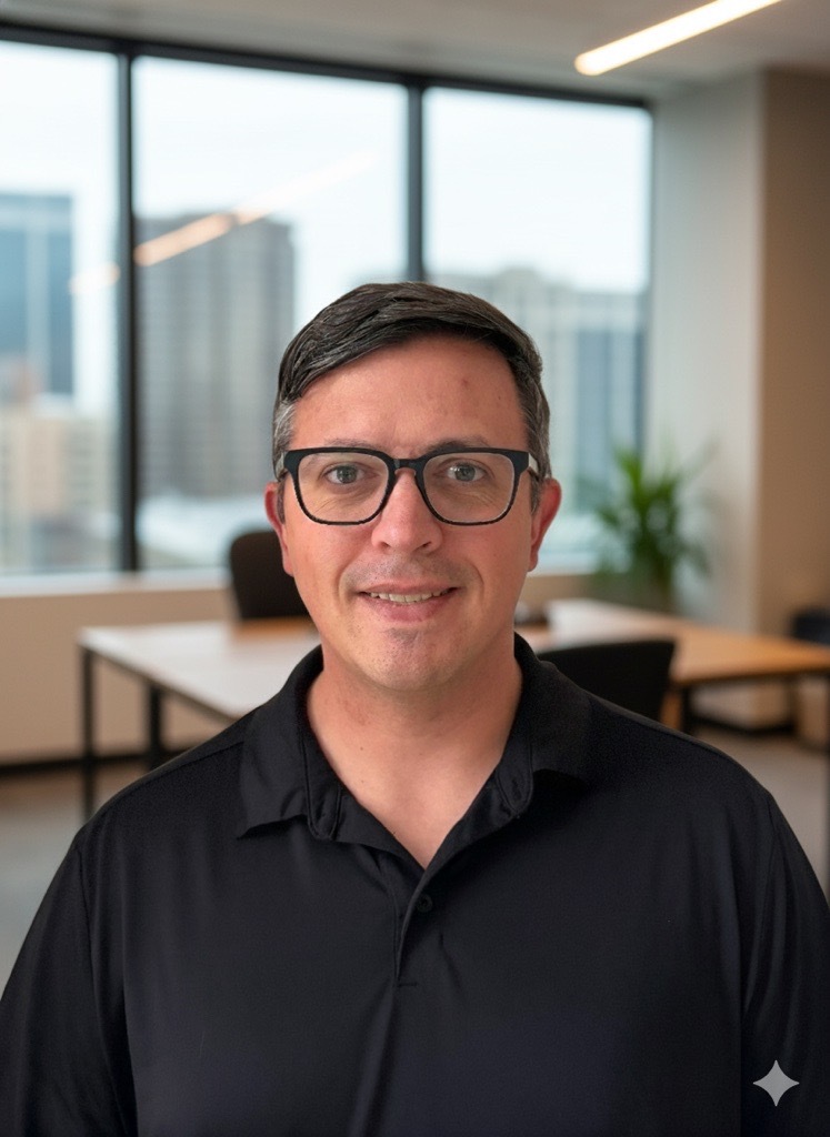 Smiling person with dark-rimmed glasses and short dark hair, wearing a black polo shirt, in a modern office setting