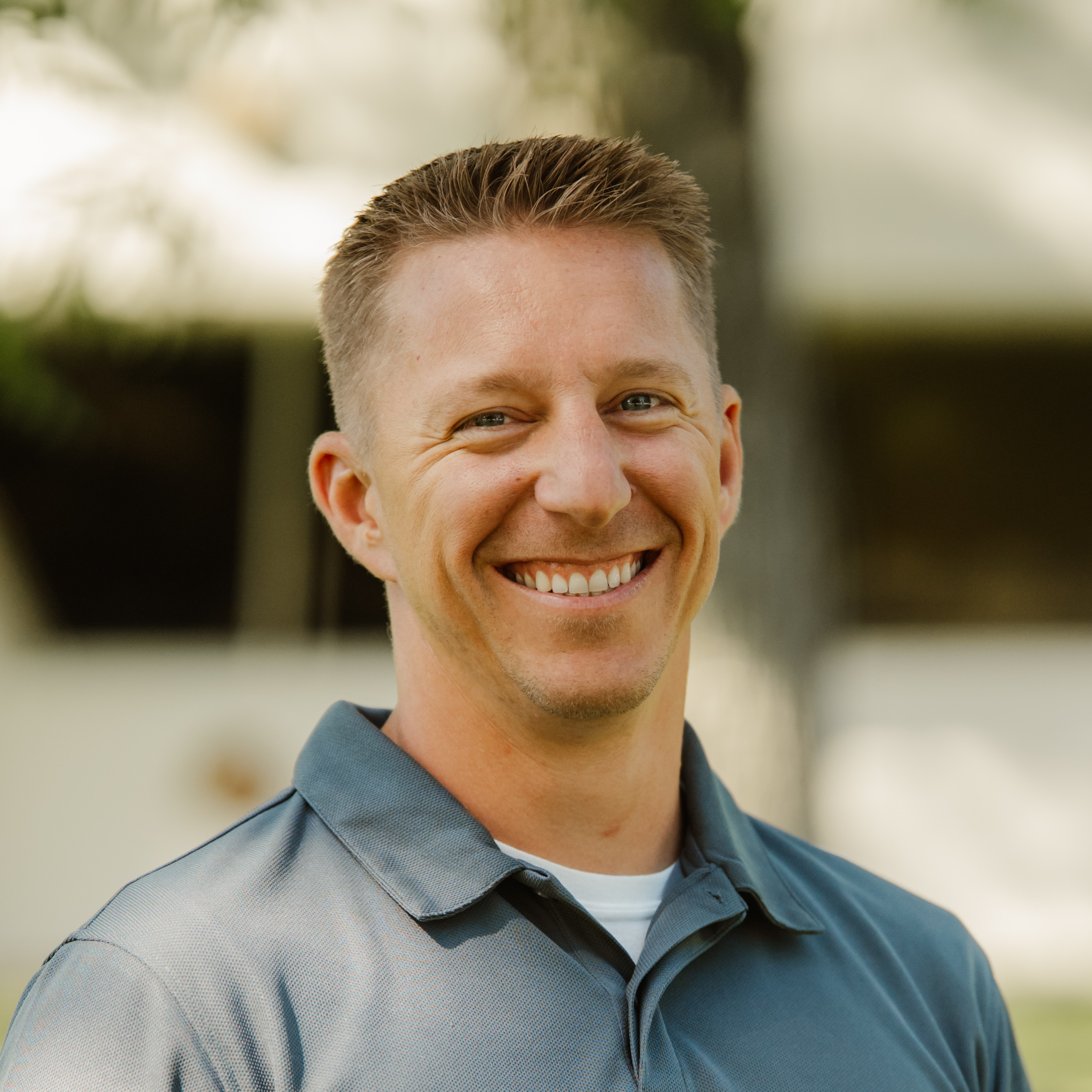 Smiling person with short hair wearing a gray polo shirt, photographed outdoors