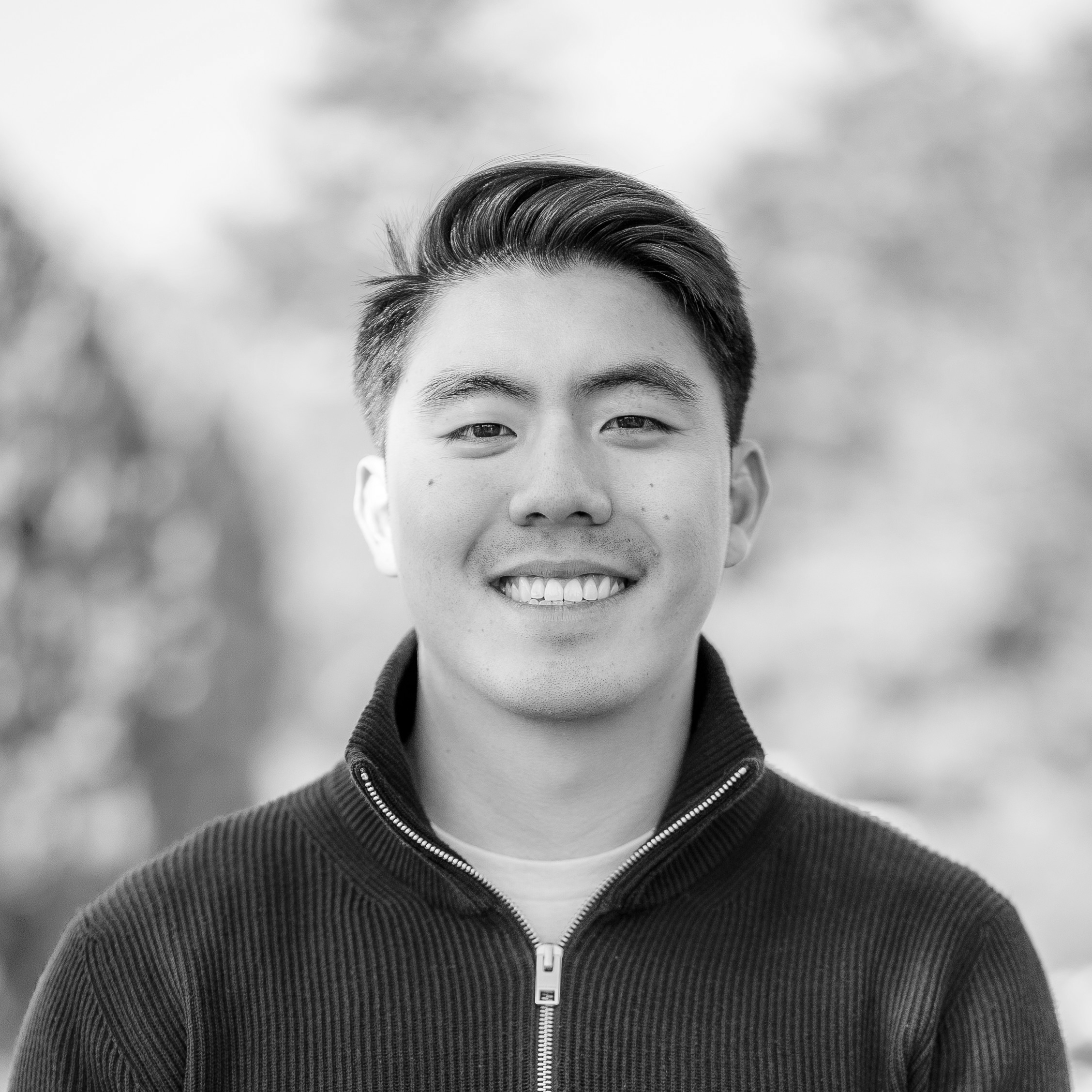 Smiling person with short dark hair wearing a dark zip-up jacket, black-and-white outdoor portrait