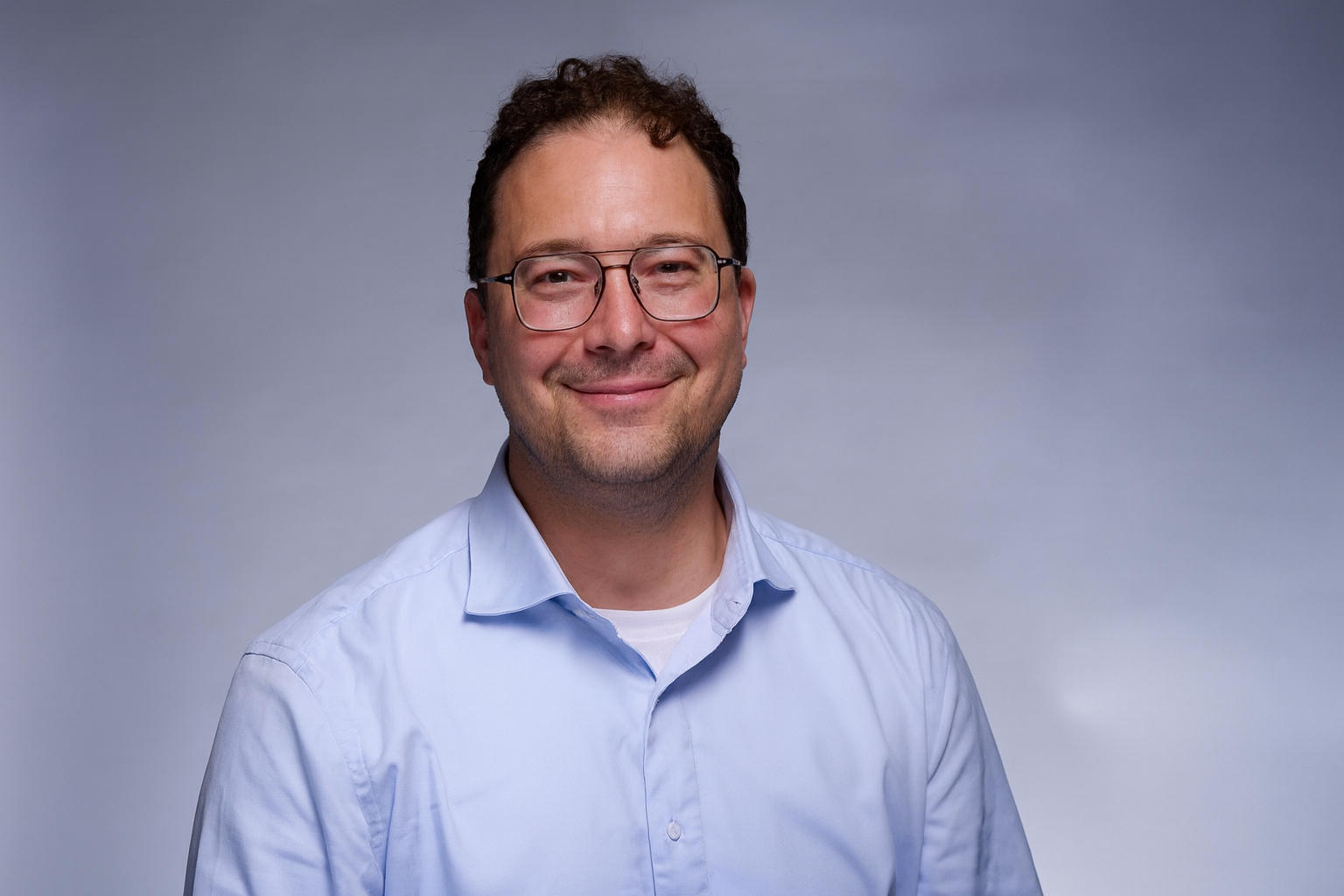Headshot of Max Ranall, smiling, wearing glasses and a light blue button-up shirt