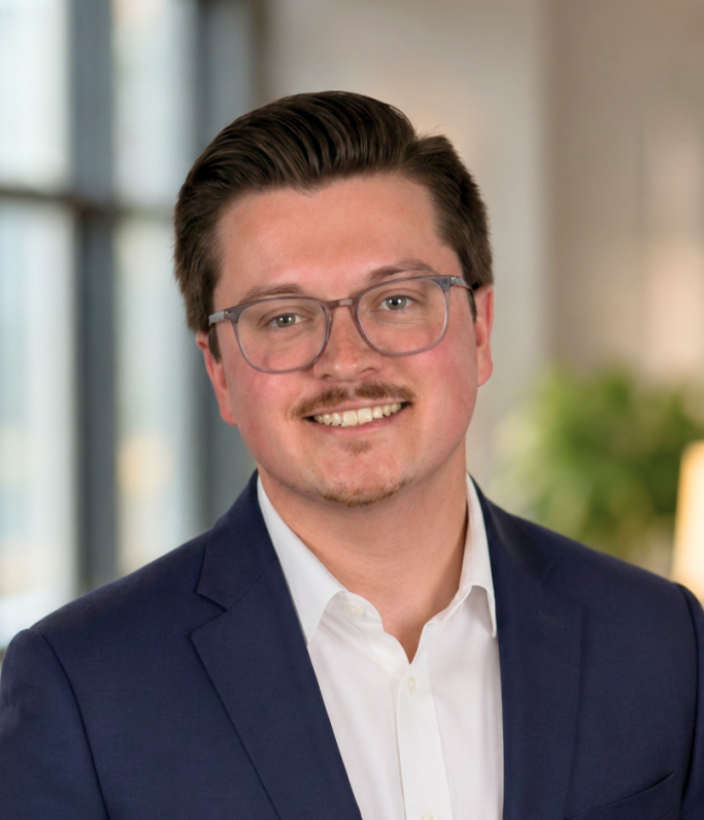 Nolan, a Hamilton Company team member, smiling in a navy blazer and glasses against a blurred indoor background