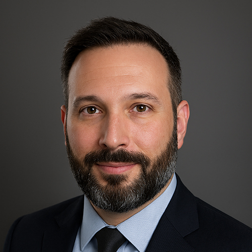 Headshot of a person with short dark hair and beard, wearing a dark suit and tie