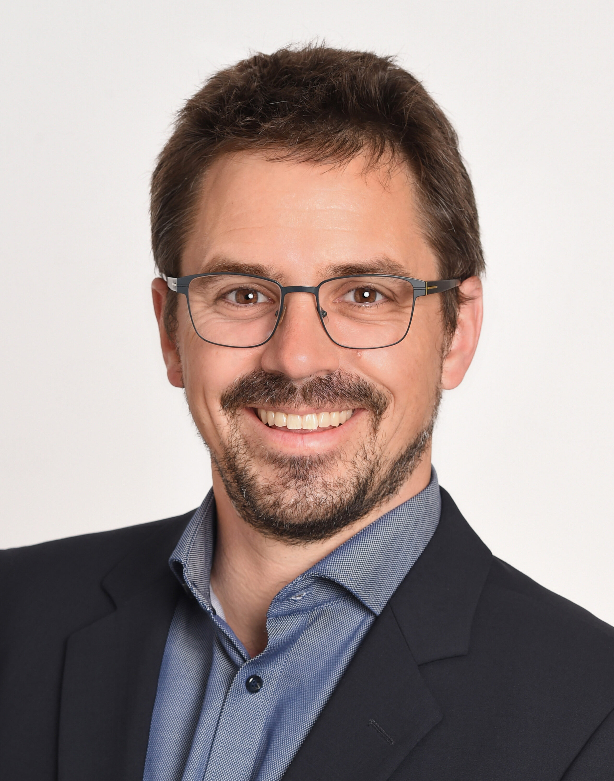 Smiling person with dark hair, beard, and glasses wearing a dark blazer and blue shirt